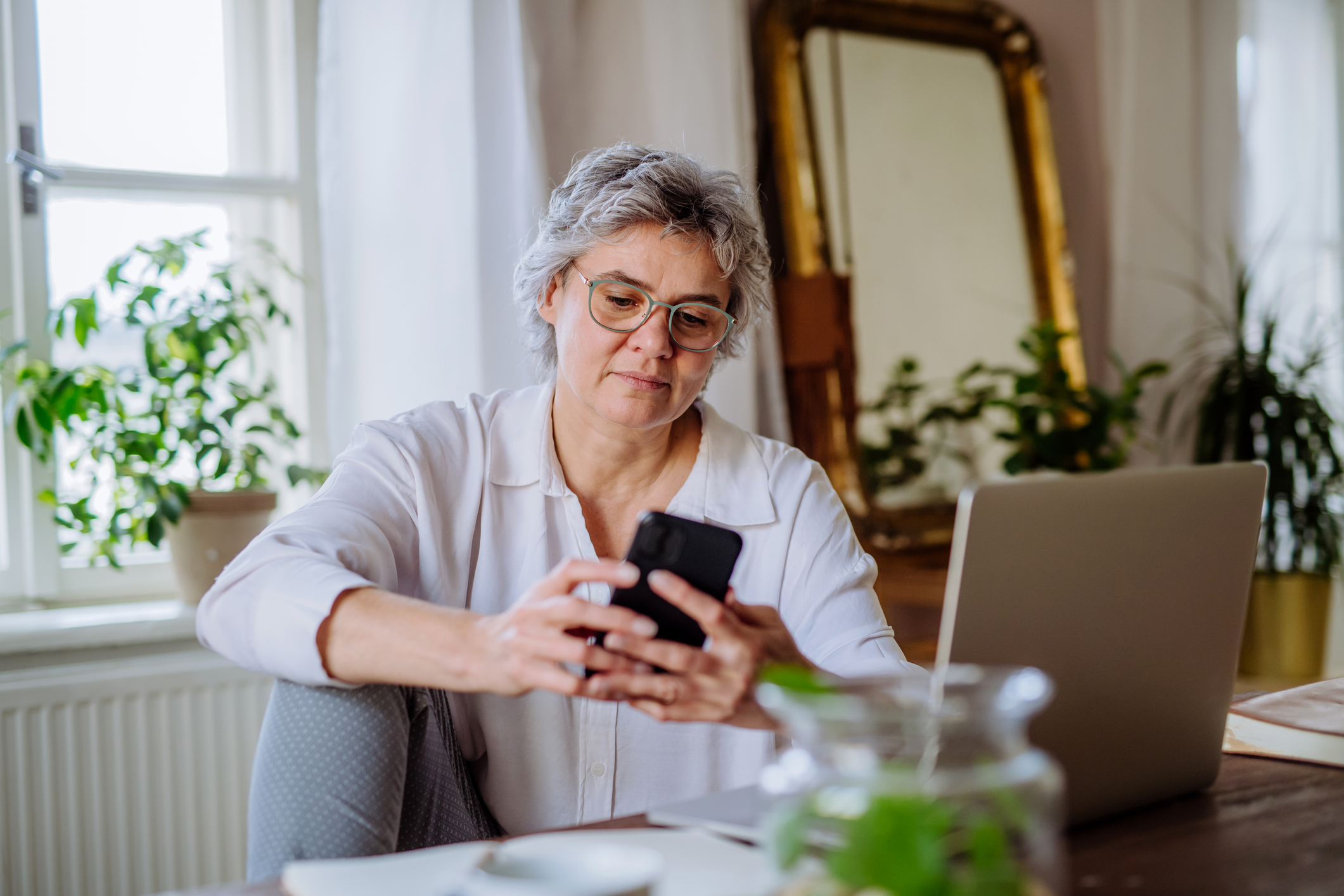 A woman at home using her smartphone and laptop simultaneously to monitor travel prices.