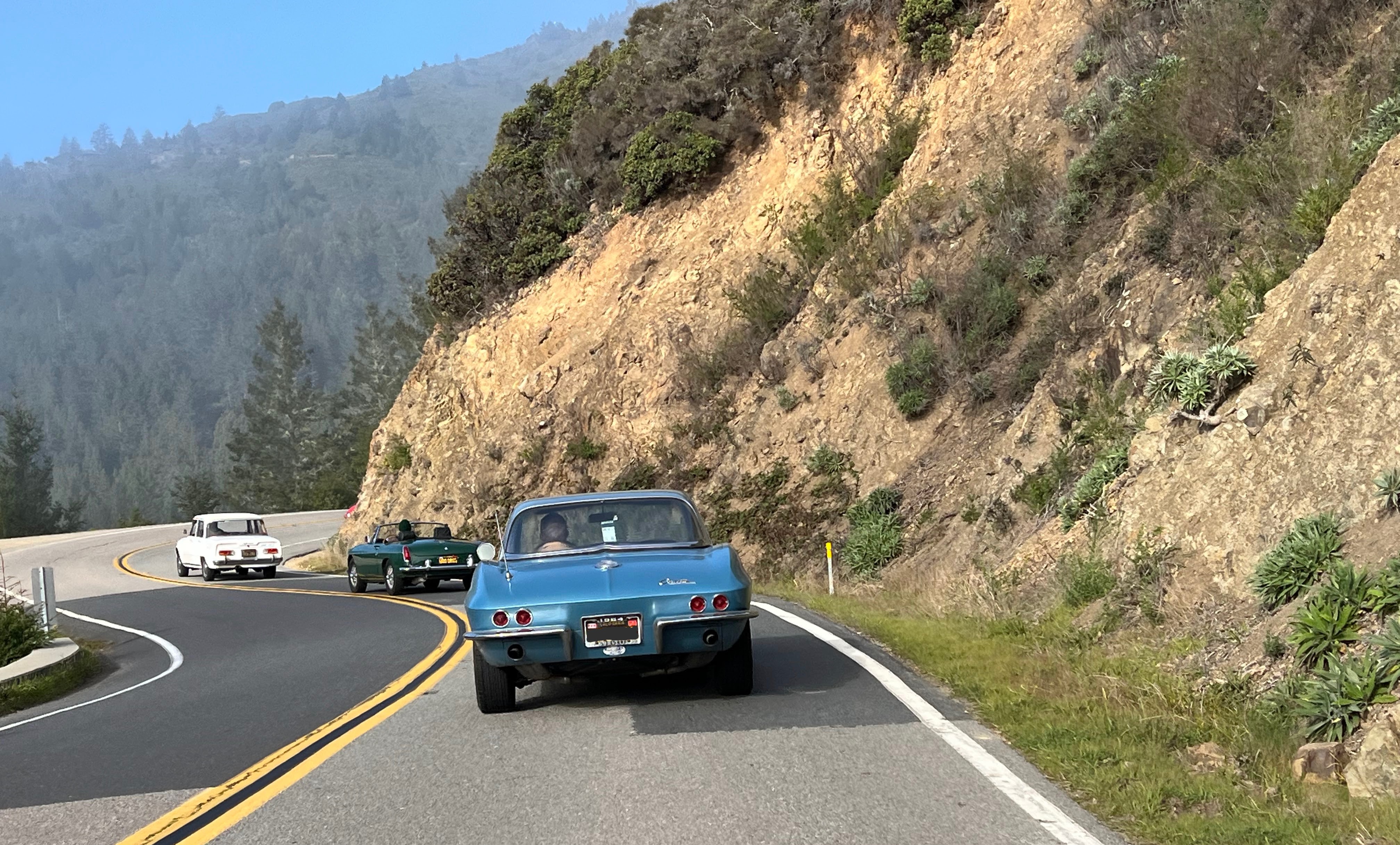 A trio of vintage sports cars, including an MG and an Alfa Romeo, driving on a winding coastal road.