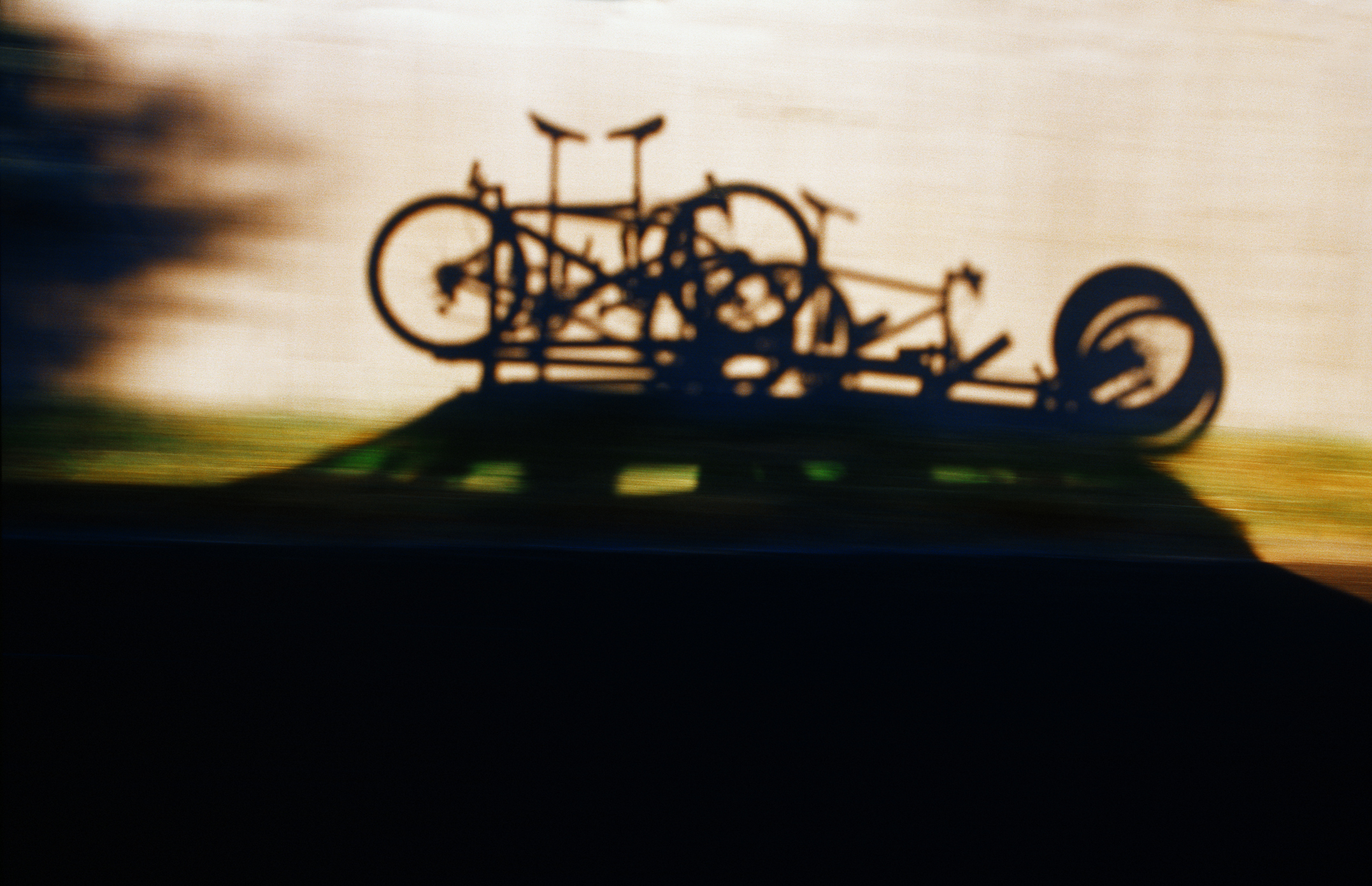Shadows of mountain bikes mounted on a vehicle's roof rack against a paved road.