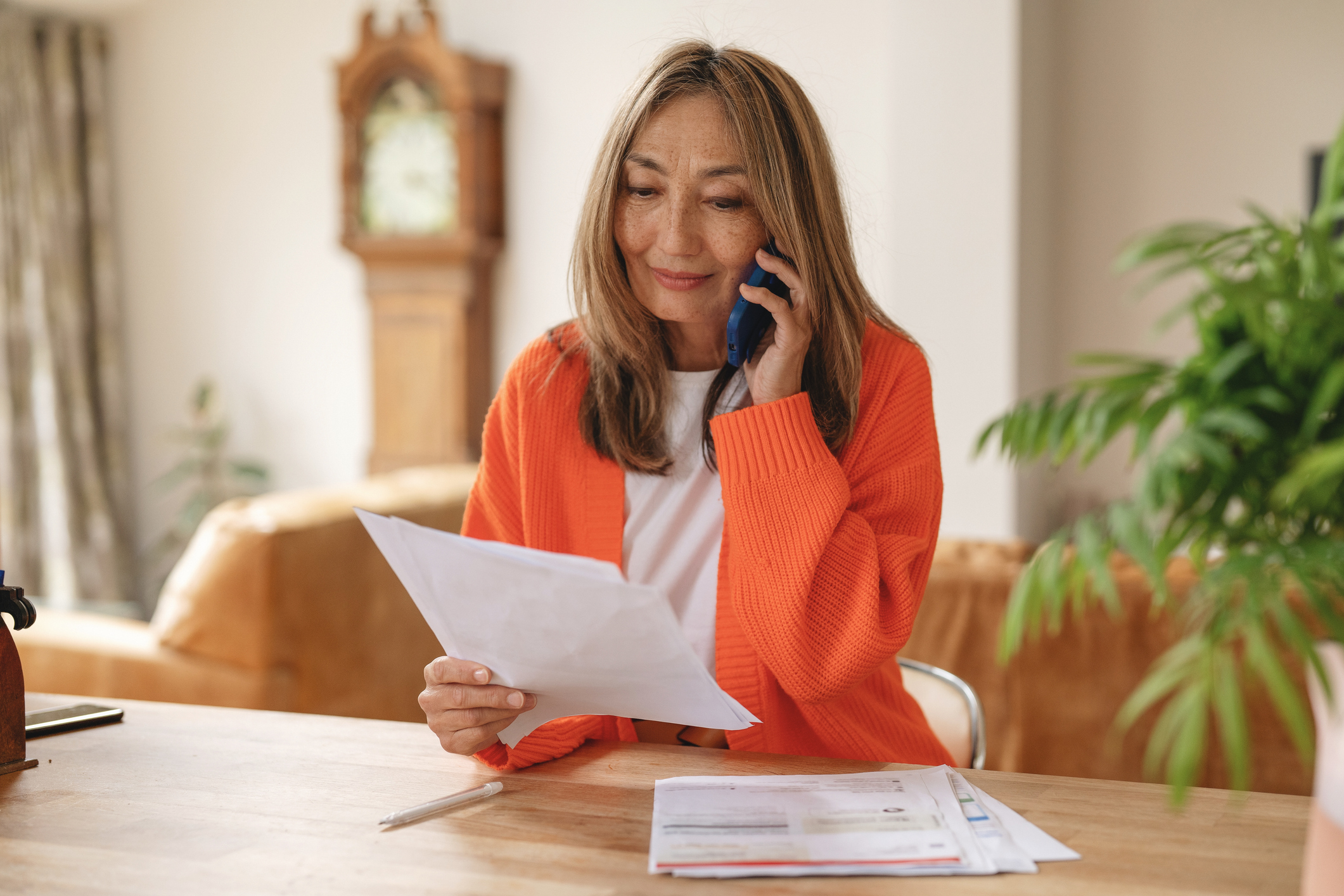 A woman sitting in a living room, talking on her smartphone while reviewing printed billing statements.