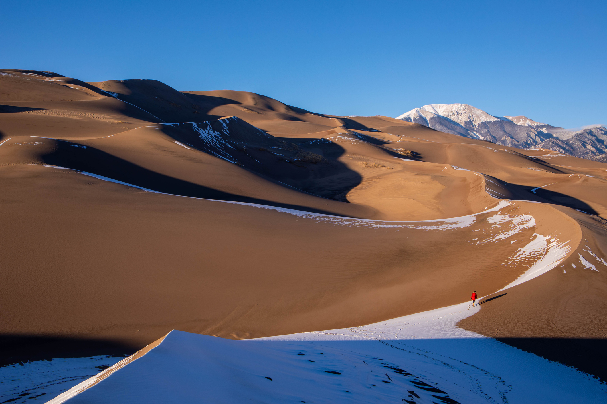 A person walking on the Great Sand Dunes with high mountain peaks in the background under bright sunlight.