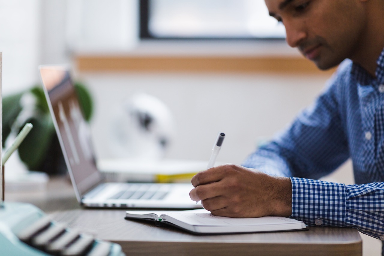 A professional checking off a detailed list on a desk with a laptop and pen.