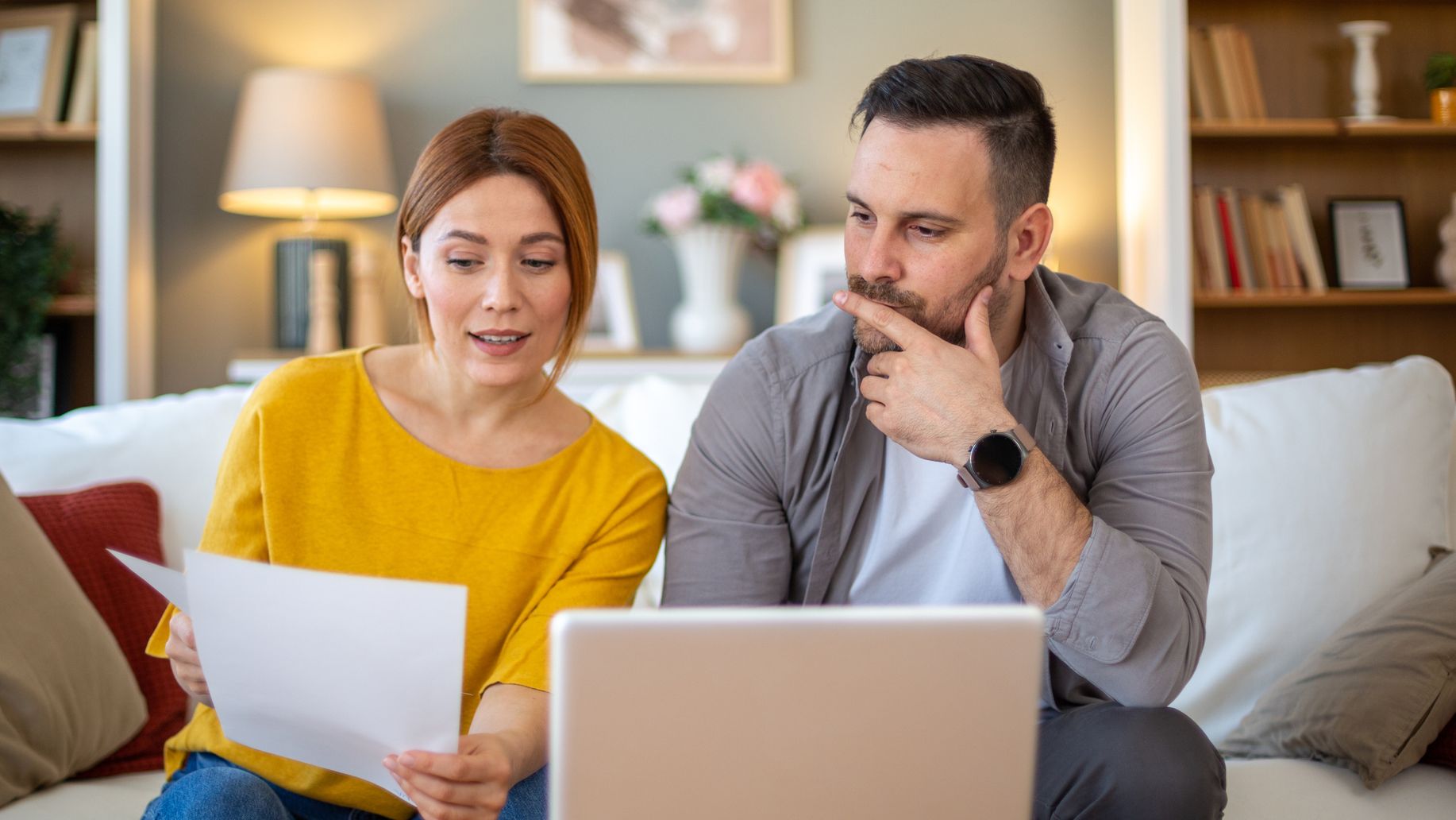 A man and woman sitting on a couch, looking at a laptop and documents while discussing their home finances.